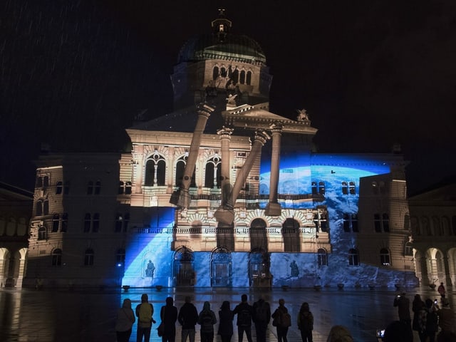 Projektion: Das Bundeshaus löst sich auf, seine Gebäudeteile fangen an zu schweben. 