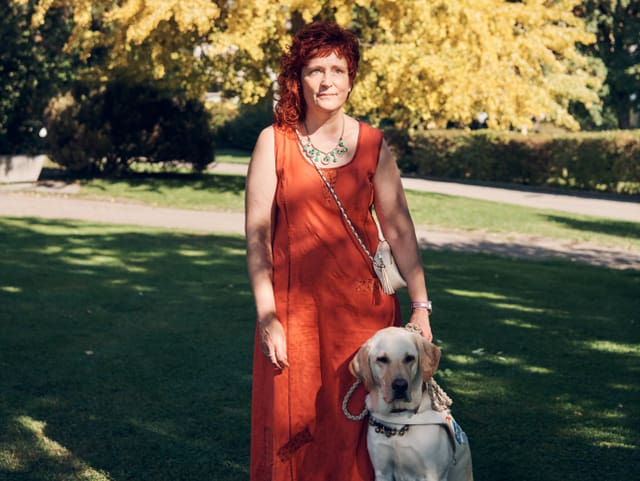 Eine Frau mit rotem Sommerkleid und roten Haaren im Grünen. Vor ihr ein sitzender Blindenhund.