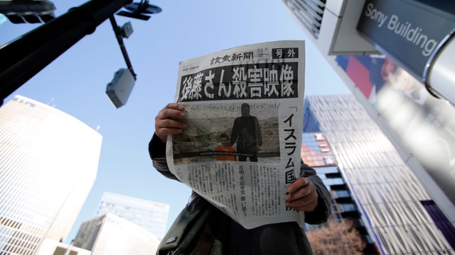 Eine Person hält zwischen Wolkenkratzern eine offensichtlich japanische Zeitung in der Hand, auf dem Titelbild ein Isis-Schlächter mit einer Geisel in orange neben sich kniend.