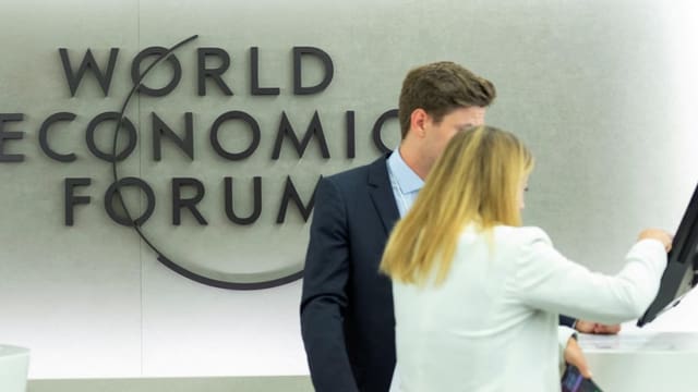 Eine Frau und ein Mann an einem Bildschirm und vor dem Logo des World Economic Forums in Davos. 