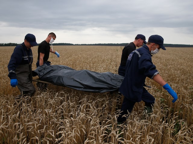 Vier Männer tragen eine in Plastik gehüllte Leiche über ein Kornfeld.