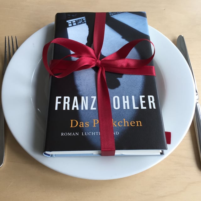 Das Buch «Das Päckchen» liegt auf einem weissen Teller mit einer Geschenkmasche drum