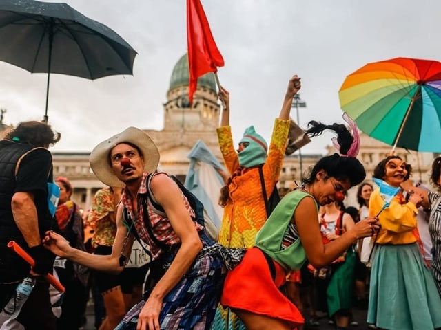 Menschen in farbenfroher Kleidung und mit regenbogenfarbigen Schirmen tanzen vor dem Nationalkongress.