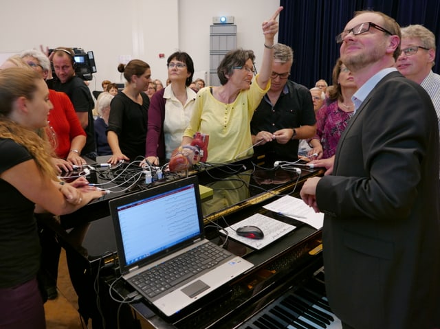 Leute stehen um ein Klavier herum, auf dem allerlei technisches Gerät steht. Eine Frau zeigt in die Höhe.