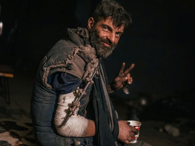 Ein verletzter Mann mit Armbinde und einer Tasse in der Hand lächelt und macht das Peace-Zeichen.