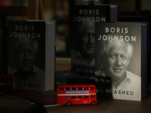 Bücherstapel mit Bild von Boris Johnson, Spielzeug-Doppeldeckerbus davor.