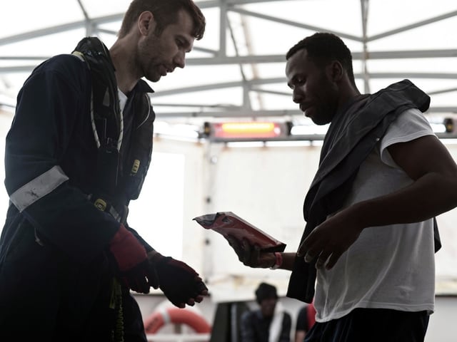 Zwei Männer, einer in Arbeitskleidung, der andere im Trainingsoutfit, unterhalten sich in einer Werkstatthalle.