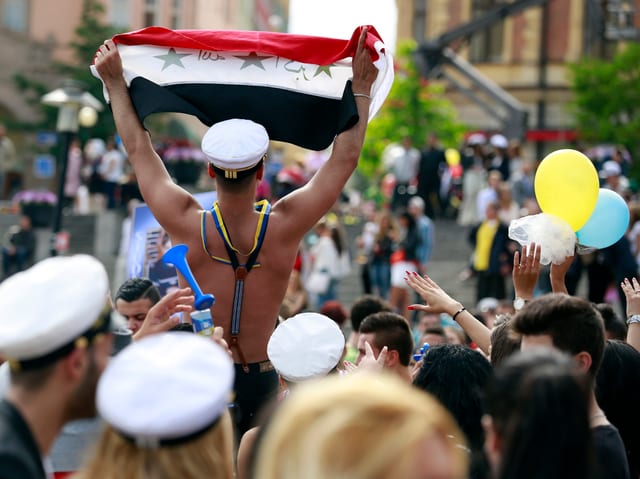 ein junger mann mit nacktem oberkörper hält an einem umzug für einen schulabschluss eine syrische flagge in die luft