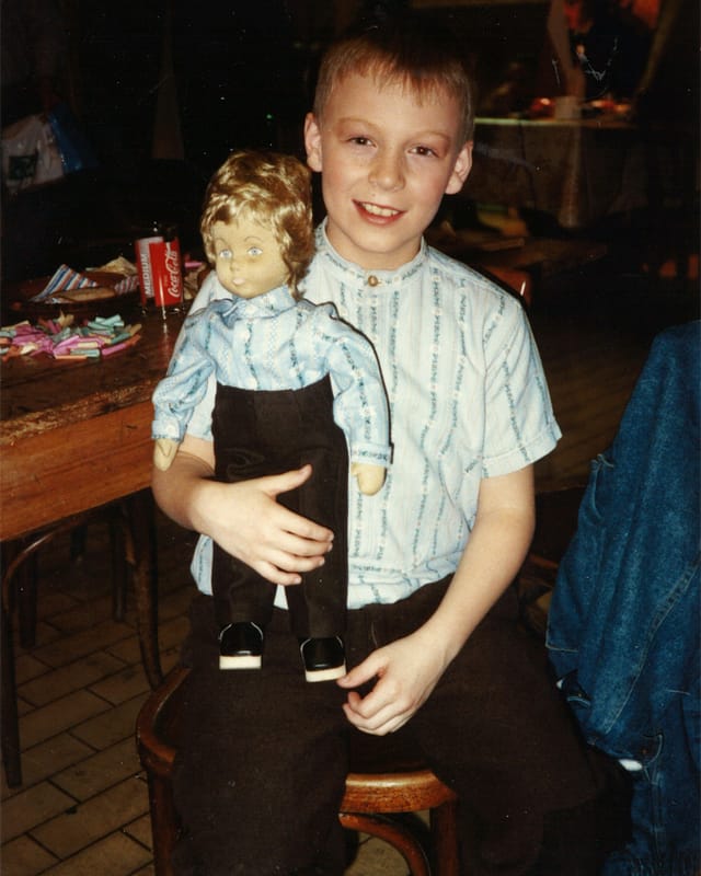 Ein blondhaariger Bub sitzt mit einer Puppe auf einem Holzstuhl.