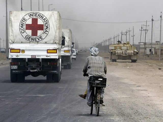 LKWs des IKRK fahren eine Strasse entlang, gefolgt von einem Mann auf einem Velo, Rechts im Bild steht ein Panzer.