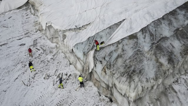 Ein weiss-grauer Berg, darauf ist sind weisse Tücher und sechs kleine Menschen in gelben Westen zu sein. 