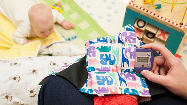 Ein Baby liegt auf dem Boden, davor hält die Mutter ein kleines Gerät in der Hand.