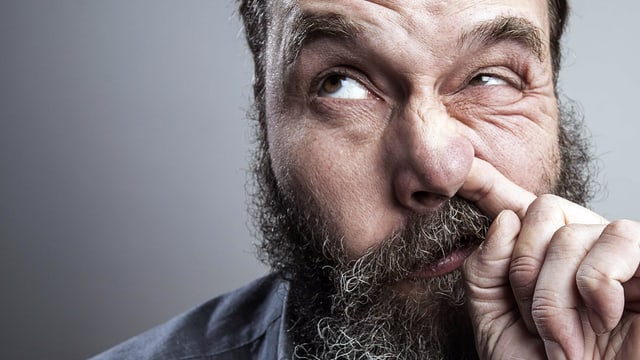 Bärtiger Mann bohrt in seiner Nase und verzieht das Gesicht dabei.