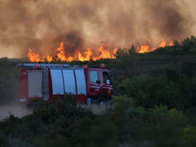 Ein Feuerwehrauto fährt den Hang hoch. Oben lodern die Flammen in den Wäldern.