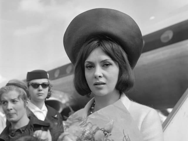 Lollobrigida mit einem Blumenstrauss in der Hand in Nahaufnahme. Hinter ihr das Flugzeug, aus dem sie gestiegen ist.