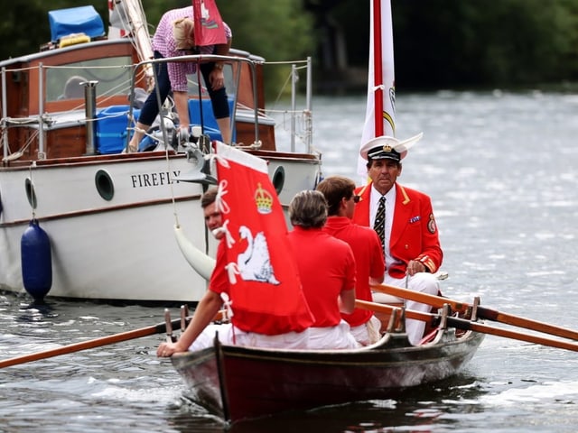 Männer in roten Blazern in einem Ruderboot auf einem Fluss nahe einem Boot.