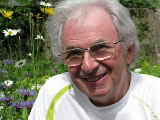 Lächelnder Mann mit Brille und grauen Haaren vor einer blühenden Wiese.