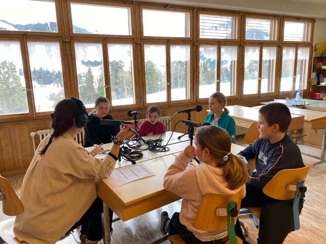 Fünf Kinder sitzen um ein Pult, auf dem sieben Mikrofone im Kreis aufgestellt sind. Die Kinder schauen Anik an.