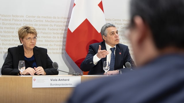 Pressekonferenz im Medienzentrum des Bundeshauses mit zwei sitzenden Personen und Schweizer Flagge im Hintergrund.