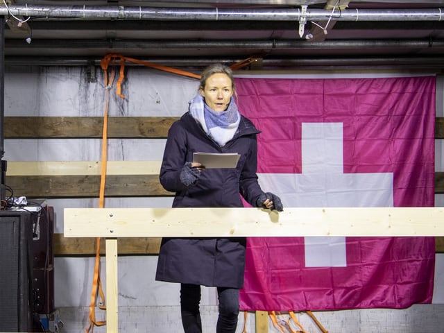 Eine Frau steht auf einer Bühne. Sie spricht zum Publikum. Sie hält einen Text in der Hand.