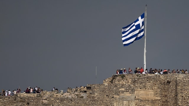 Symbolbild: Griechische Flagge aus der Ferne flattert im Wind, darunter Menschen. 