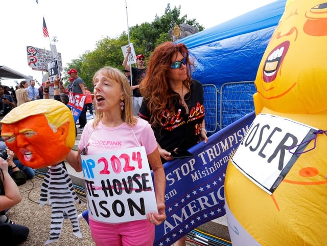 Eine Frau hält eine Trumpmaske, daneben ist jemand in einem Trump-Babykostüm zu sehen.