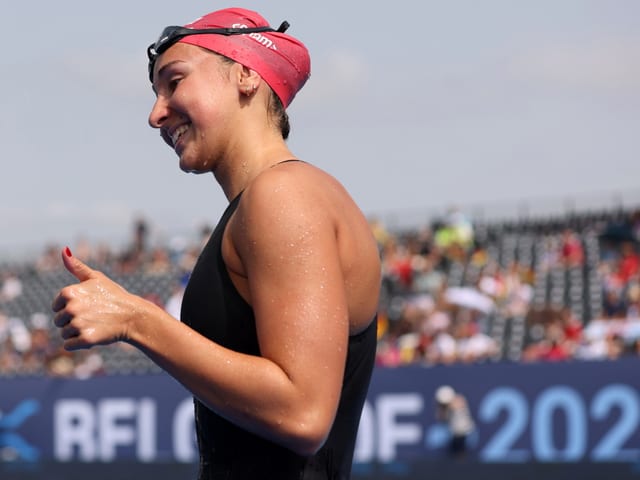 Schwimmerin mit roter Badekappe gibt Daumen hoch bei Schwimmwettbewerb.