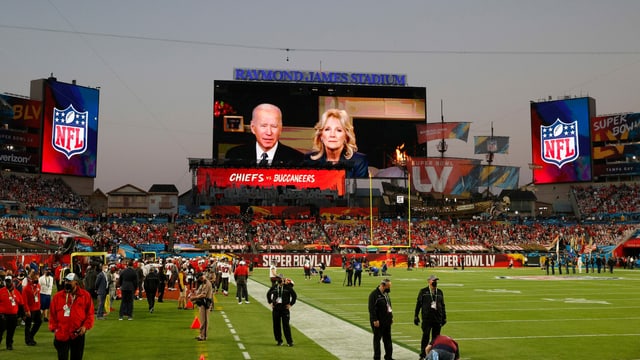 A soccer stadium with people and a video message of Joe and Jill Biden playing in the background. 