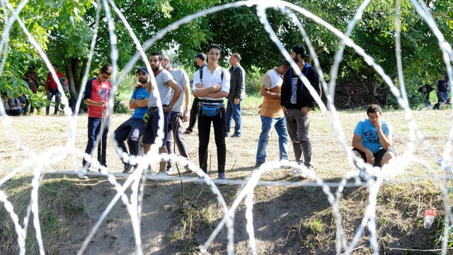 Hinter einem Stacheldrahtzaun stehen und sitzen Flüchtlinge.