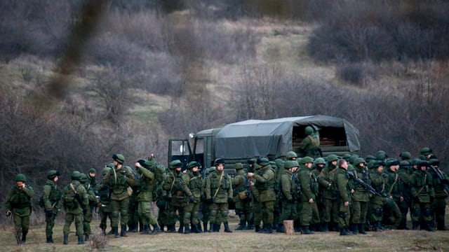 Mehrere Dutzend bewaffnete Soldaten in grünen Uniformen sammeln sich in der Ferne um einen Truppentransporter.