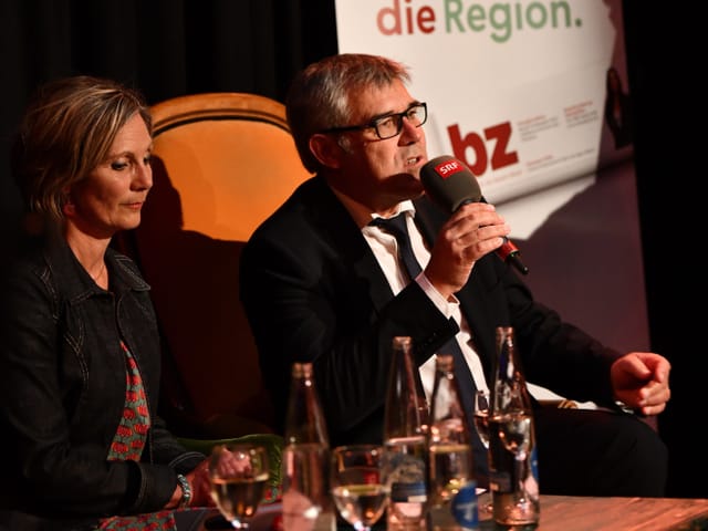 Zwei Personen sitzen auf einem Sofa. Eine Person hält ein Mikrophon in der Hand und spricht. 