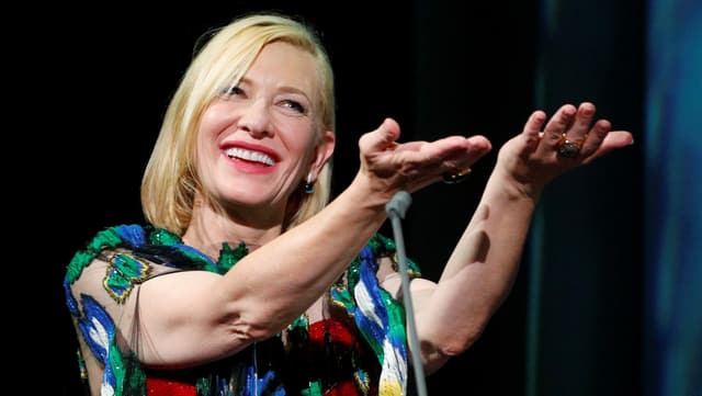 Kate Blanchett smiles at the camera and extends both hands forward.