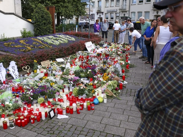 Menschen stehen vor einem Gedenkstätte mit Blumen und Kerzen.