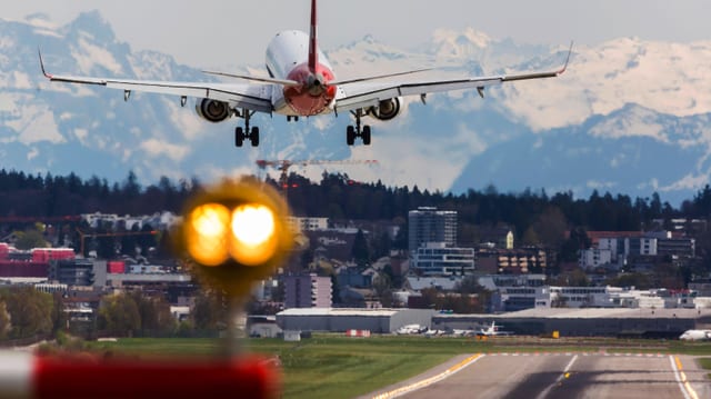 Ein Flugzeug fliegt über der Landebahn, im Hintergrund sind schneebedeckte Alpen zu sehen.