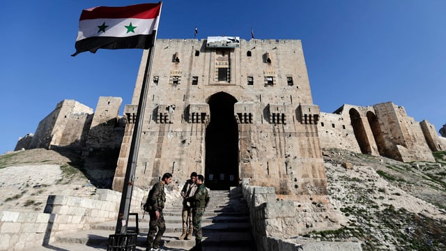 Soldaten vor einem Gebäude. An einem Fahnenmast weht die syrische Flagge.