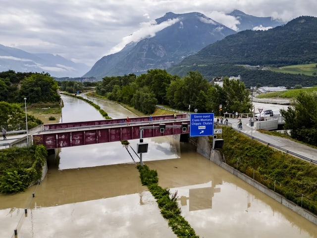 Überflutete Strasse unter einer Brücke, mit Bergen im Hintergrund.