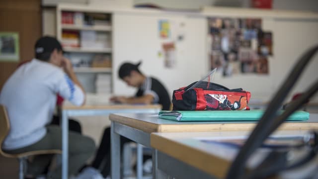 Vorne scharf ein Etui mit Geodreieck, dahinter verschwommen zwei jugendliche Schüler mit Kappe