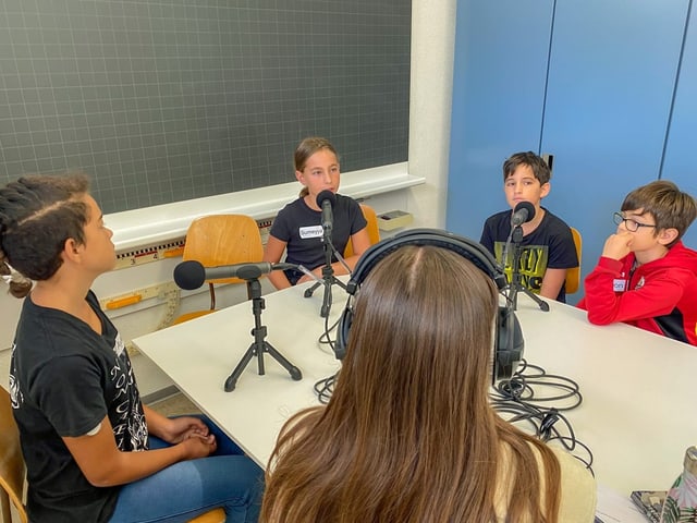 Kinder sitzen an einem Tisch und sprechen in Mikrofone.