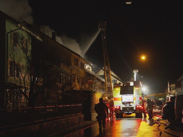 Ein Feuerwehrwagen steht in einer Strasse. Ein Feuerwehrkran ist ausgefahren, von dem aus ein Brand bekämpft wird.