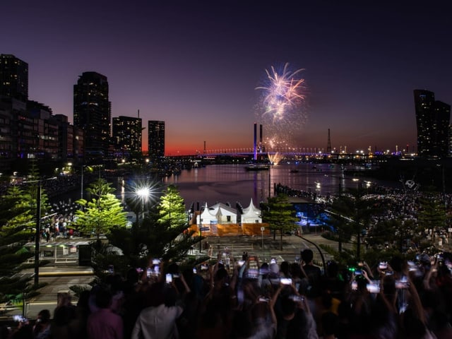 Feuerwerk leuchtet über dem Hafenbecken in Melbourne. Im Vordergrund stehen Zuschauende.