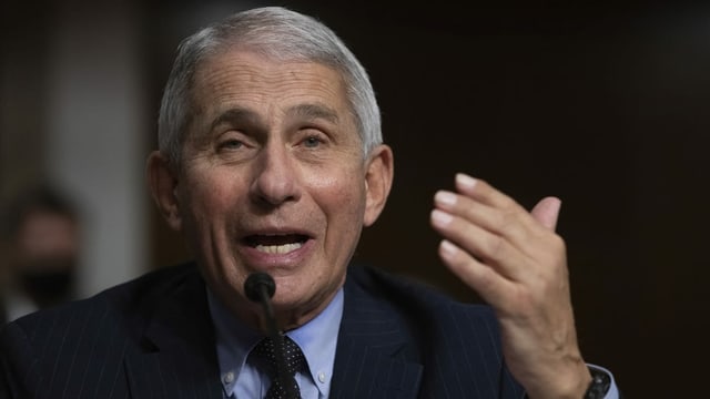 Fauci speaks into a microphone and gestures with one hand.