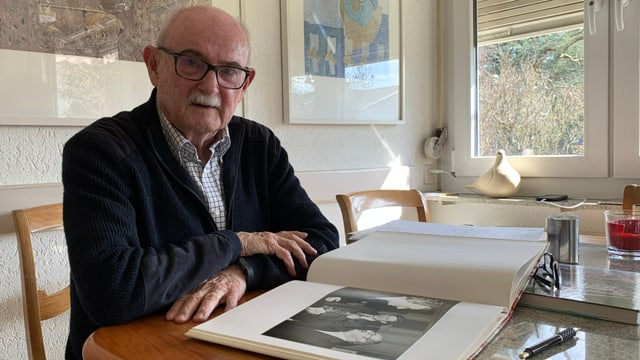 Älterer Mann mit Brille sitzt an Tisch, hat Fotobuch vor sich.