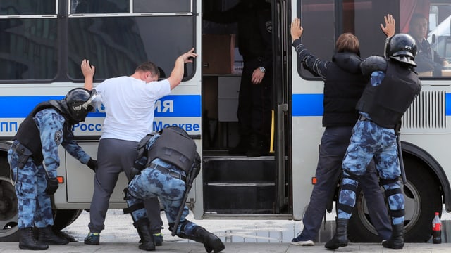 Polizeibus. Davor werden Personen von Polizisten durchsucht. Sie stehen mit erhobenen Armen und gespreizten Beinen vor dem Bus.