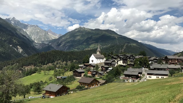 Ein kleines Dorf mit alten Holzhäusern und einer Kirche umgeben von Bergen und Wäldern.