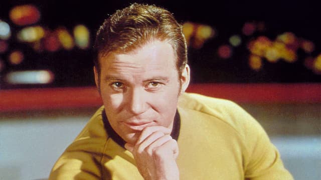 Shatner als Kirk in gelbem Pulli in Raumschiffkulisse – er blickt lächelnd in die Kamera