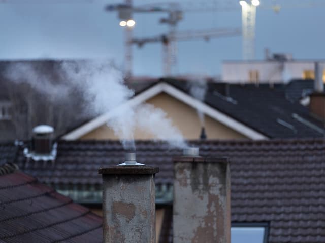 Rauchender Schornstein vor einer städtischen Kulisse mit Kränen.