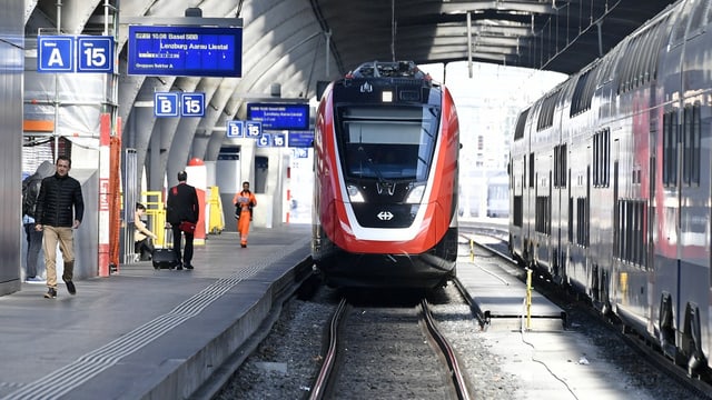 rot-weiss glänzender Zug fährt auf Gleis ein, links Perron mit Leuten.