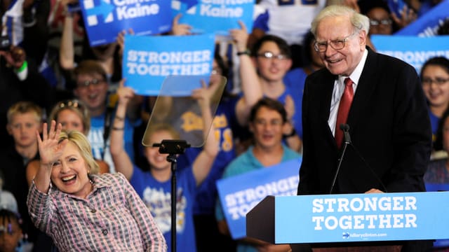 Warren Buffett steht an einem Rednerpult; Hillary Clinton daneben winkt.