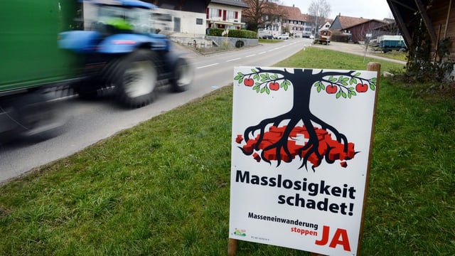 Ein Traktor mit Anhänger braust an einem SVP-Plakat (Masslosigkeit schadet!) auf einer Wiese vorbei, hinten ein Dorf.