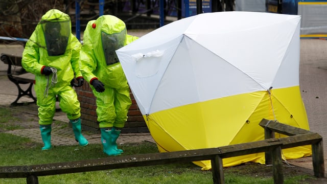 Zwei Polizisten in gelben Schutzanzügen nähern sich einem weiss-gelben Zelt.